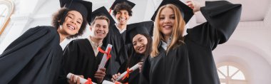 Low angle view of smiling interracial graduates holding diplomas, banner  clipart