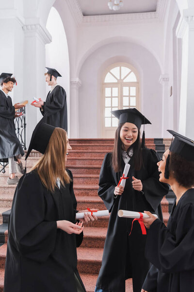 Smiling multiethnic students with diplomas talking in university 