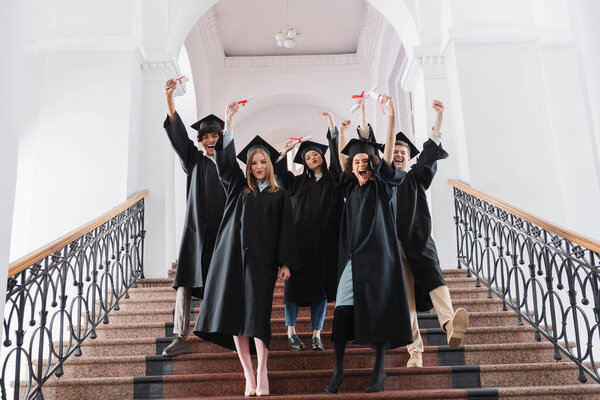Excited multiethnic graduates with diplomas standing on stairs in university hall