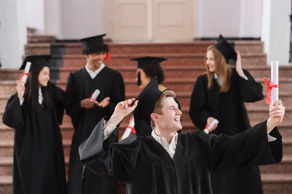 Soltero Sonriendo Mientras Mira Diploma Cerca Amigos Interracial Fondo Borroso —  Fotos de Stock