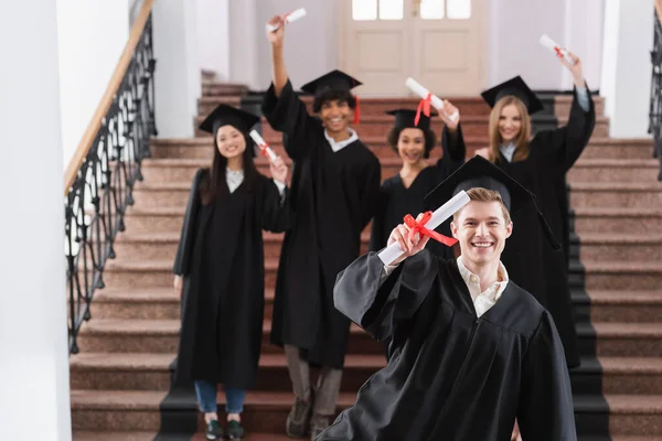 Bachelor with diploma smiling near interracial students on blurred background