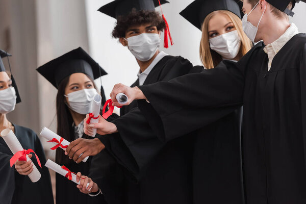 Multicultural bachelors in medical masks on blurred background holding diplomas 