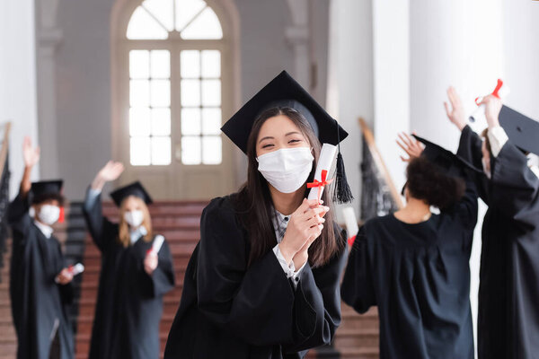 Asian bachelor in cap and medical mask holding diploma near blurred friends 