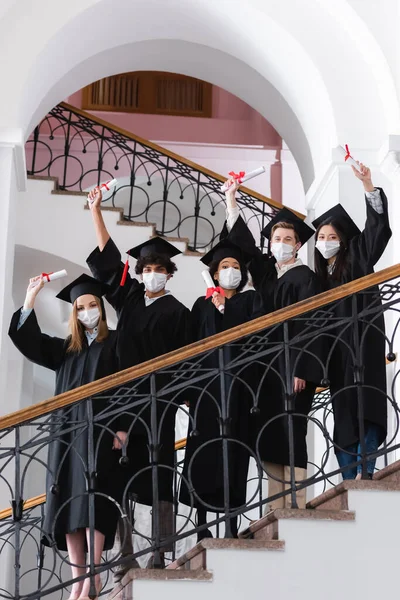 Multicultural students in medical masks and academic caps holding diplomas on stairs in university