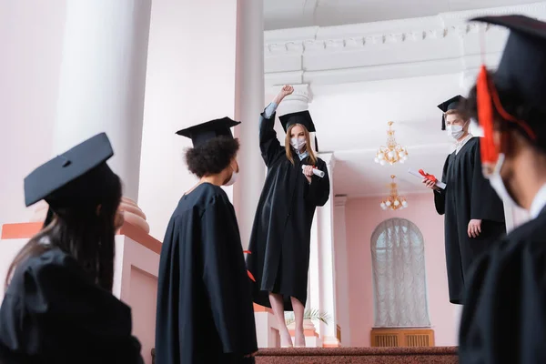 Licenciado Máscara Médica Con Diploma Mostrando Gesto Cerca Amigos Multiétnicos —  Fotos de Stock