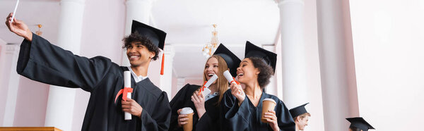 Smiling multiethnic graduates with paper cups taking selfie, banner 