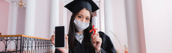 Asian graduate in medical mask holding smartphone with blank screen and diploma, banner 