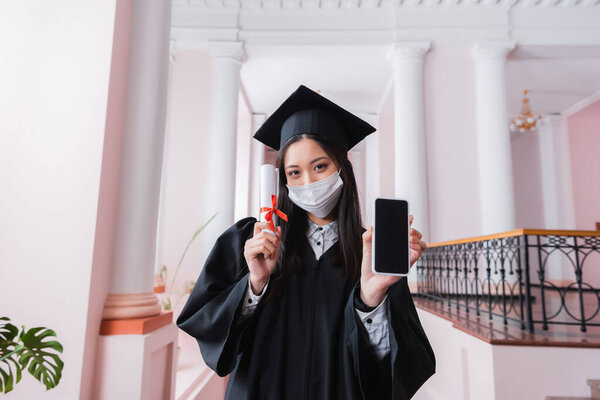 Asian bachelor in medical mask holding diploma and smartphone in university 