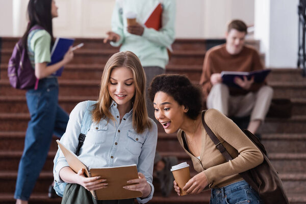 Excited african american student with coffee standing near friend with notebook 