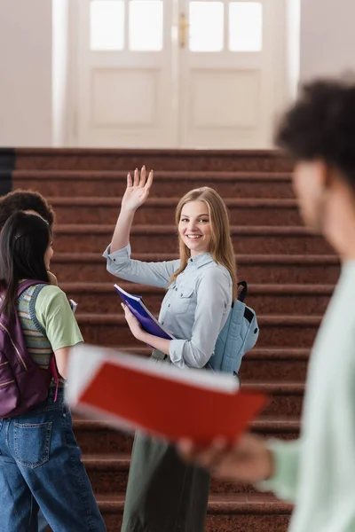 Uśmiechnięty Student Notatnikiem Machający Ręką Niewyraźnego Przyjaciela — Zdjęcie stockowe