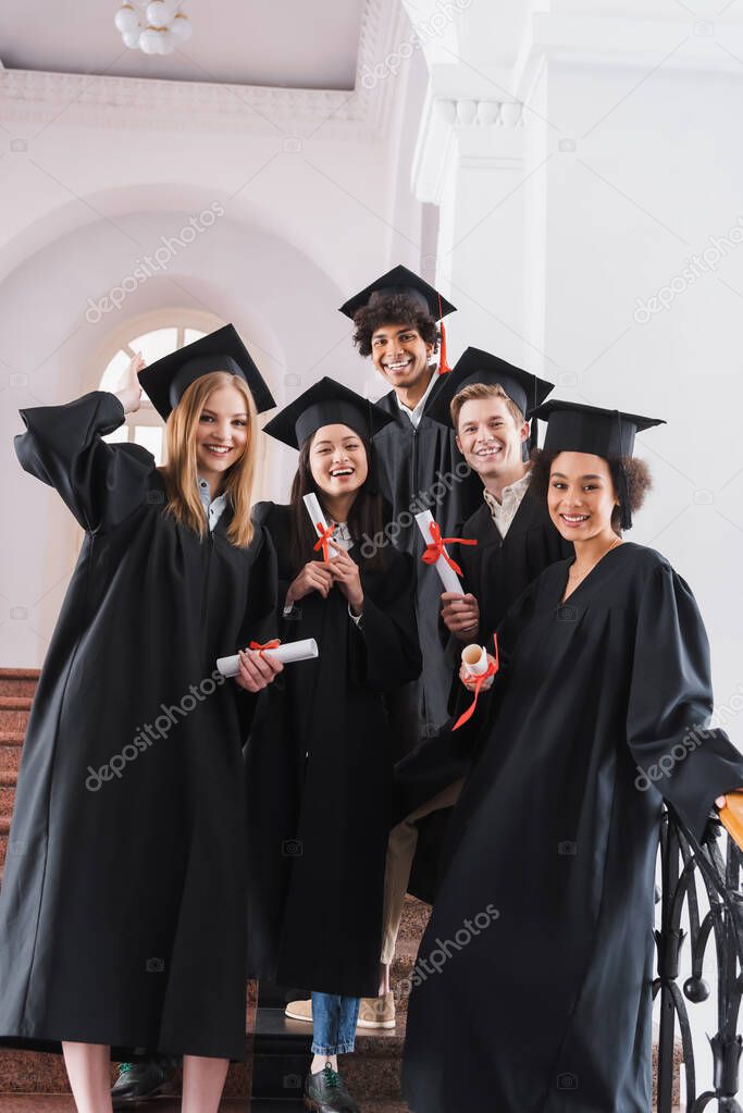 Smiling multicultural graduates in academic dresses holding diplomas