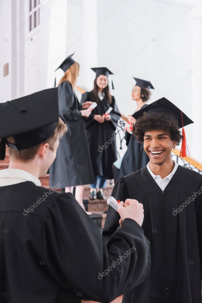 Smiling african american graduate with diploma standing near friend on blurred foreground 