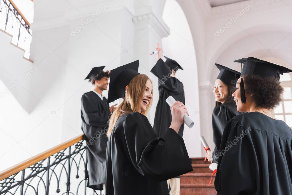 Smiling graduate in cap holding diploma near interracial friends in university 