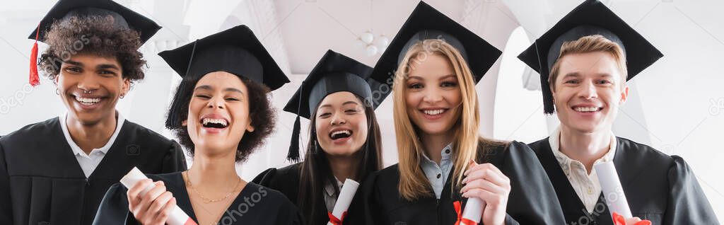 Multiethnic students in caps holding diplomas and smiling at camera, banner 