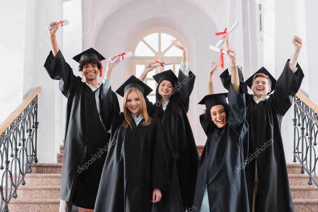 Cheerful multicultural graduates holding diplomas and showing yes gesture 