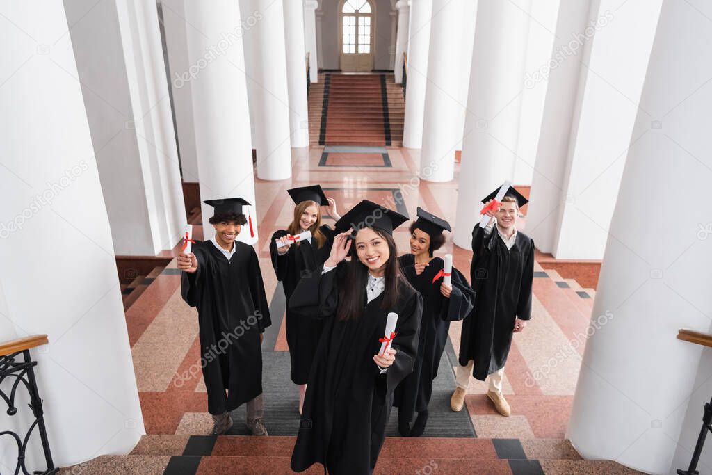 High angle view of cheerful asian graduate holding diploma near friends in caps 