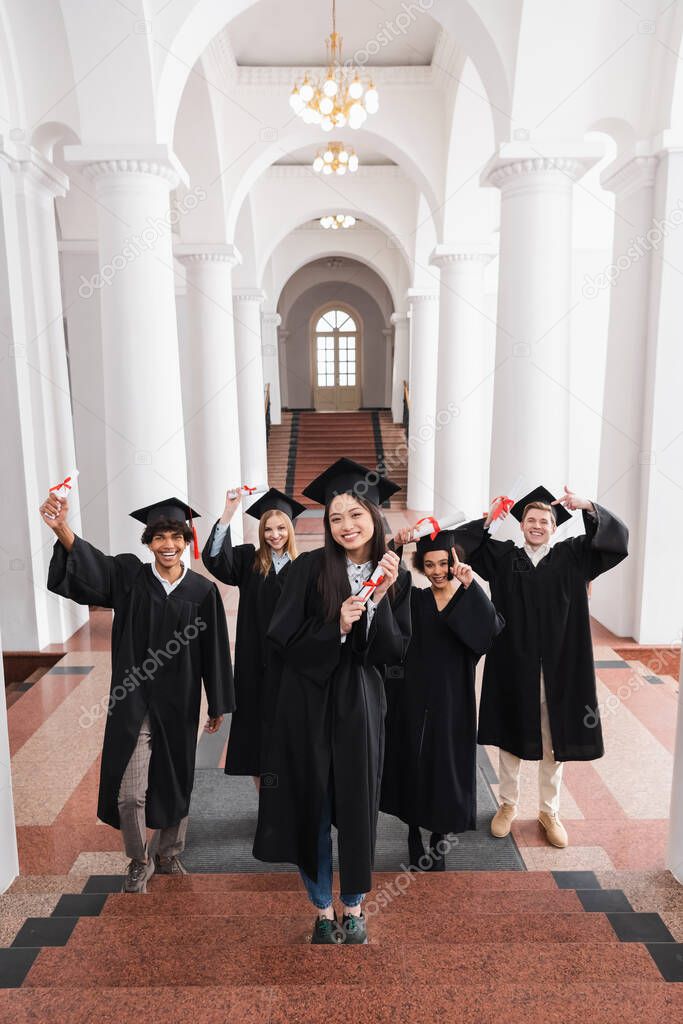 Smiling asian graduate holding diploma on stairs near friends in university 