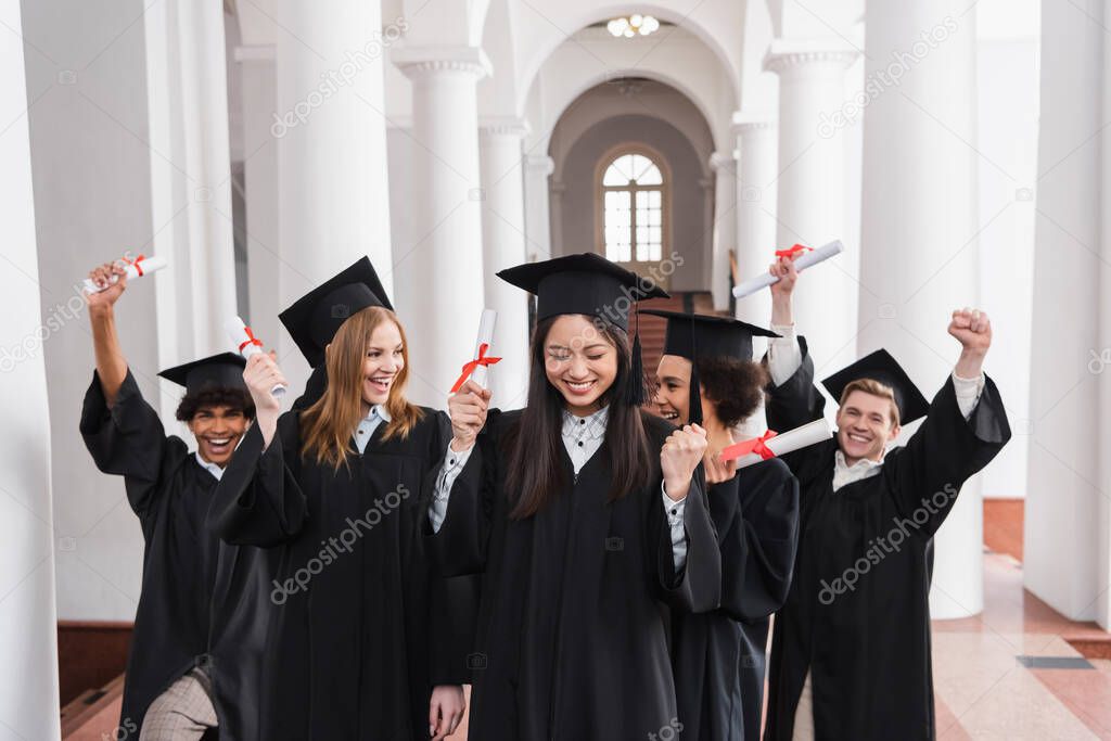 Asian graduate with diploma showing yes gesture near excited friends 