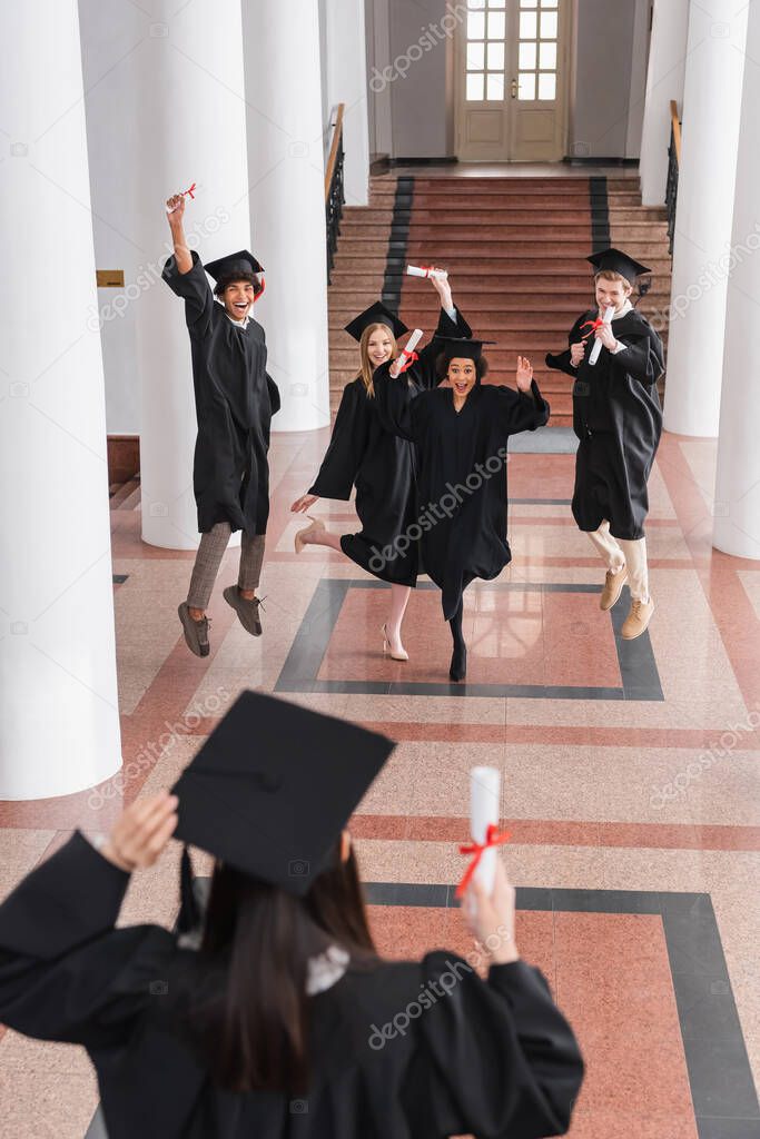 Excited multiethnic graduates with diplomas jumping near friend on blurred foreground