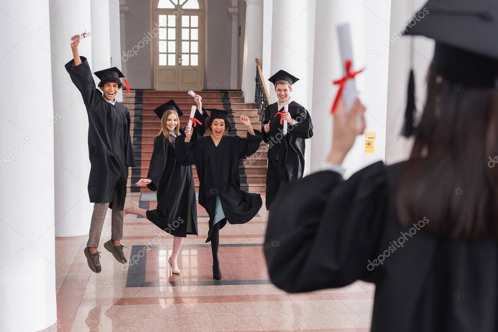 Cheerful multiethnic graduates holding diplomas near friend on blurred foreground 