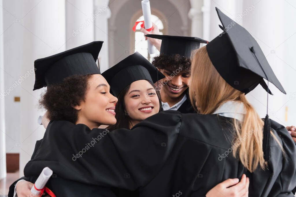 Cheerful asian graduate looking at camera while hugging friends 