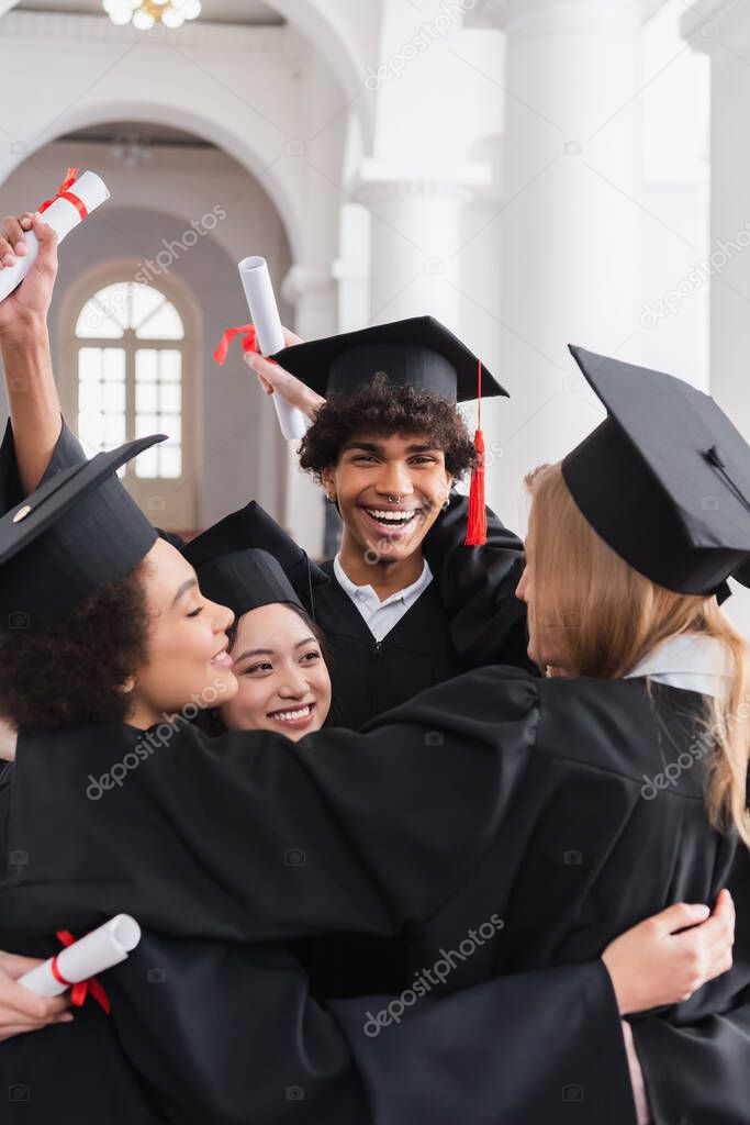 Cheerful multiethnic students with diplomas embracing in university 