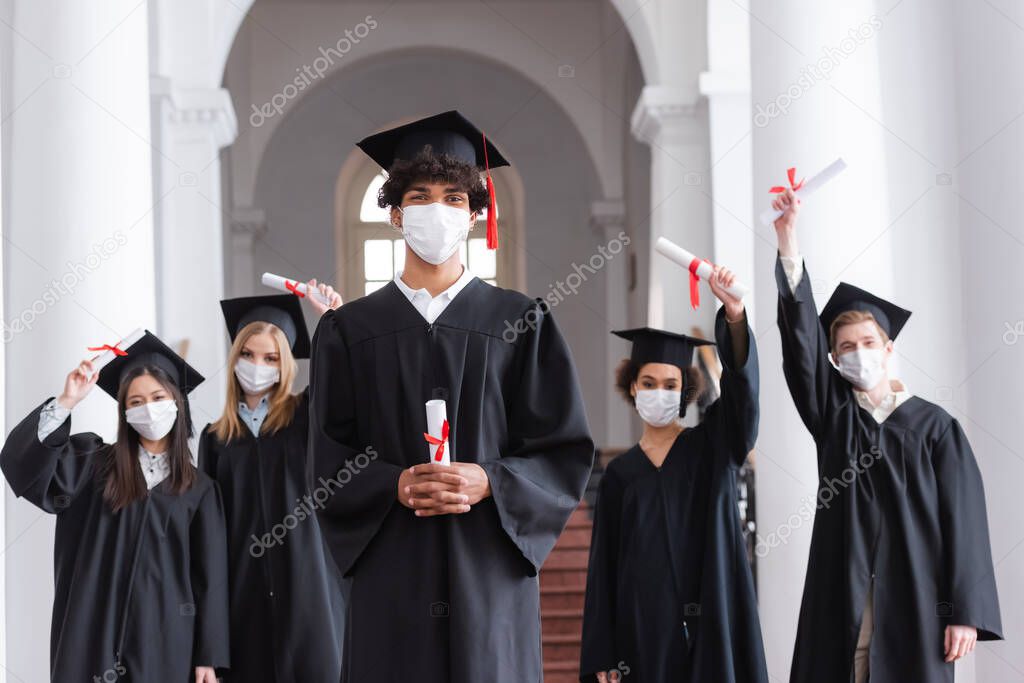 African american graduate in medical mask holding diploma near friends on blurred background 