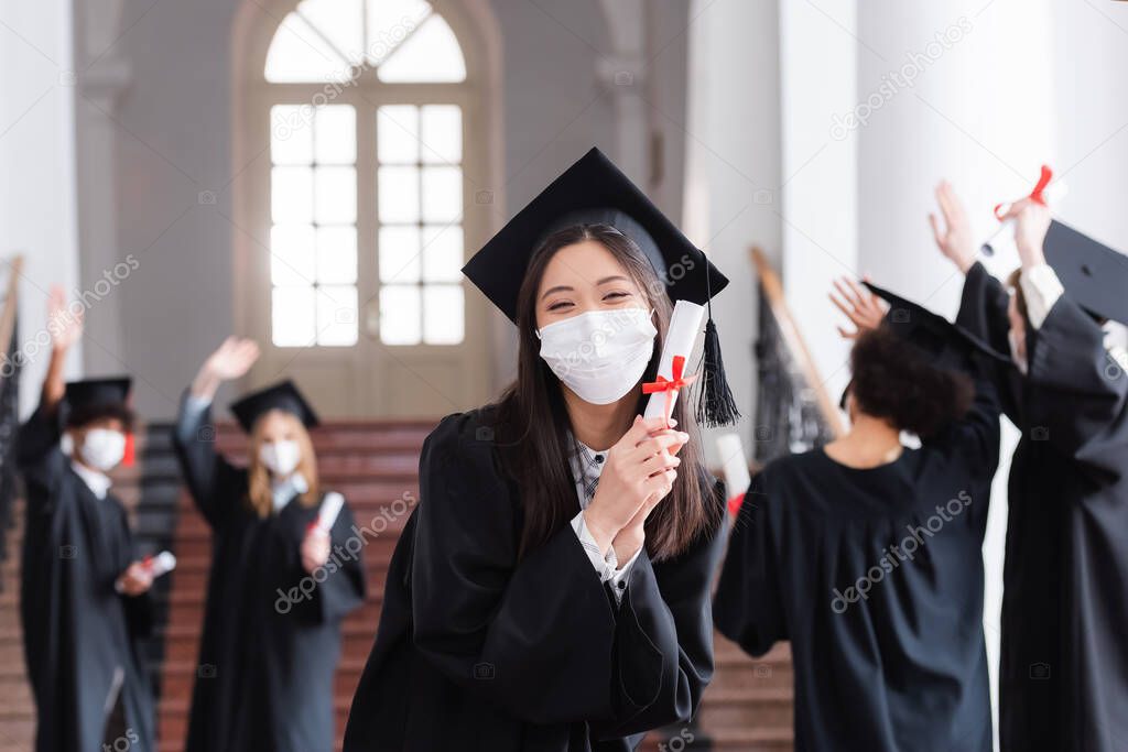 Asian bachelor in cap and medical mask holding diploma near blurred friends 