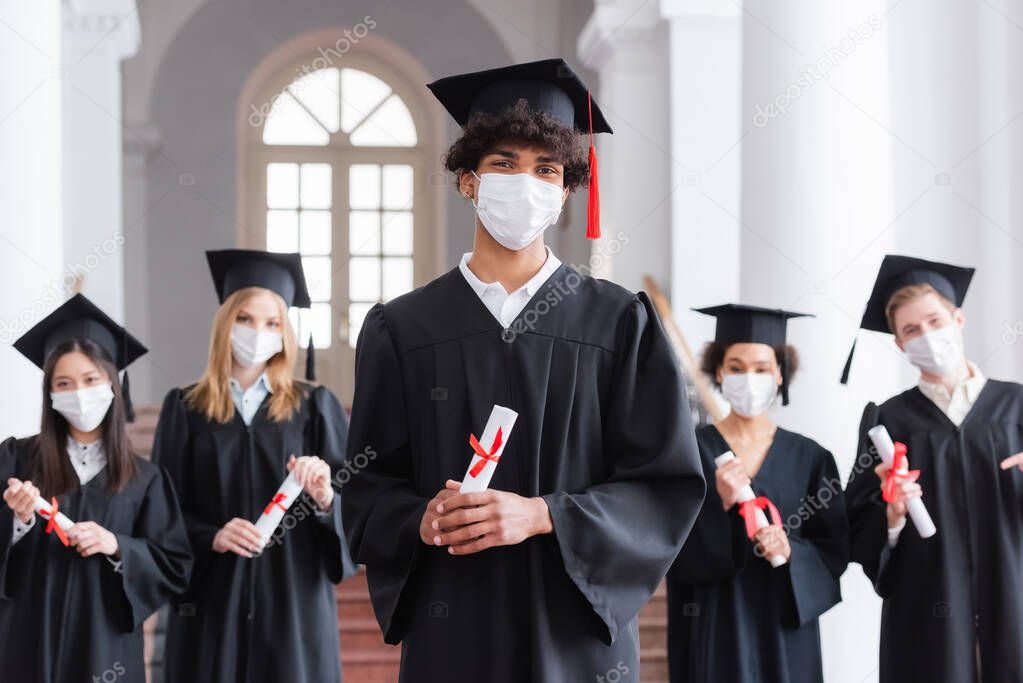 African american graduate in medical mask and academic dress holding diploma 