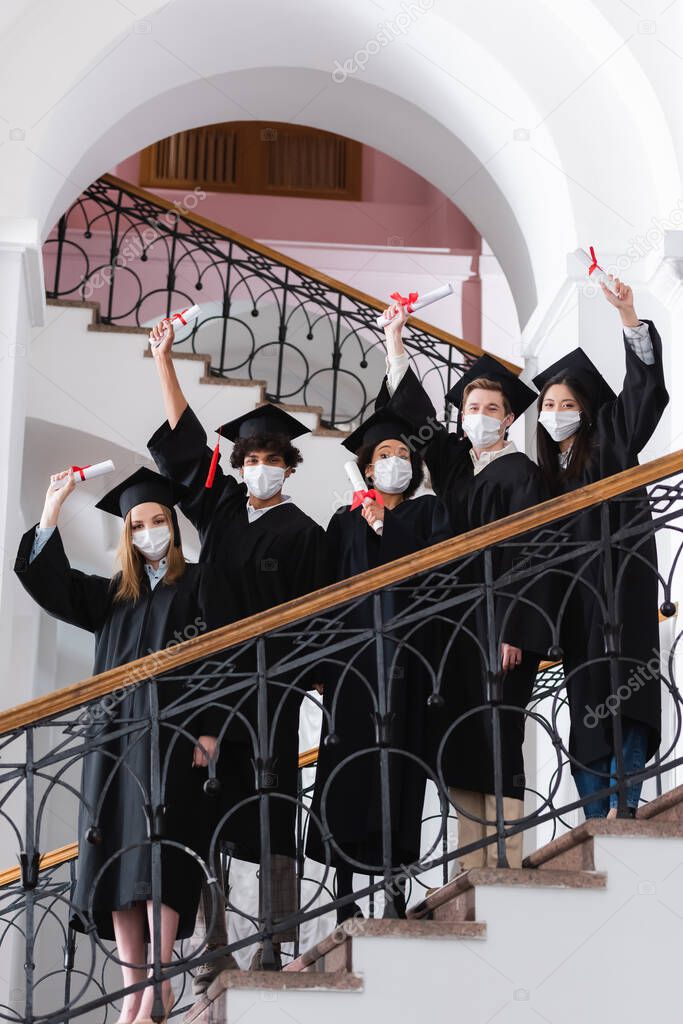 Multicultural students in medical masks and academic caps holding diplomas on stairs in university 