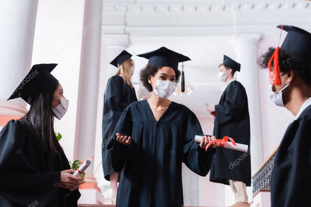 African american graduate holding diploma near friends in medical masks on blurred foreground 