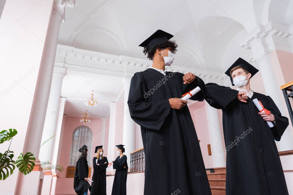 Low angle view of multiethnic graduates in medical masks doing elbow bump in university 