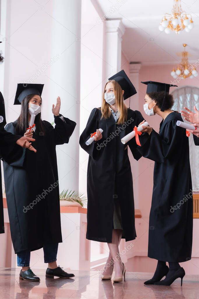 Multiethnic graduates in medical masks holding diplomas and talking in hall of university 