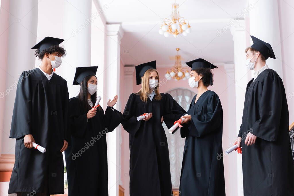 Multicultural graduates in protective masks holding diplomas and talking 