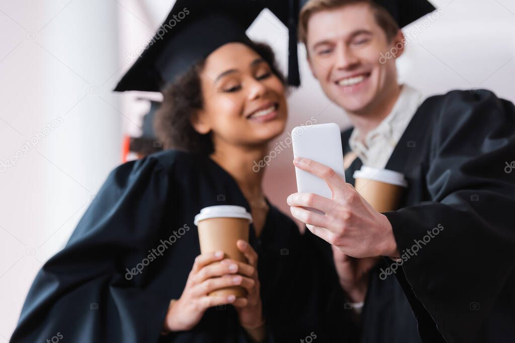 Smartphone in hand of graduate on blurred background near african american friend with paper cup 