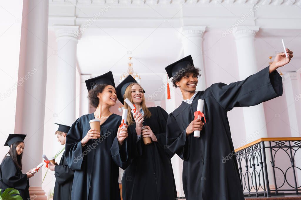 Cheerful multiethnic graduates with diplomas and paper cups taking selfie 