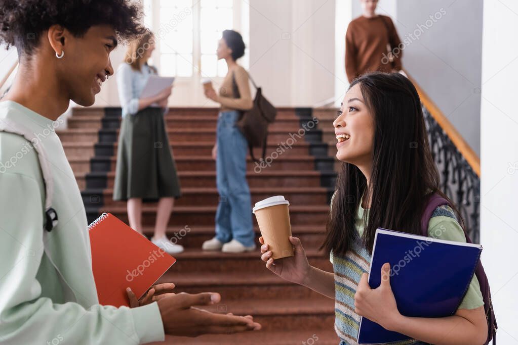 Asian student with takeaway drink talking with african american friend with notebook 