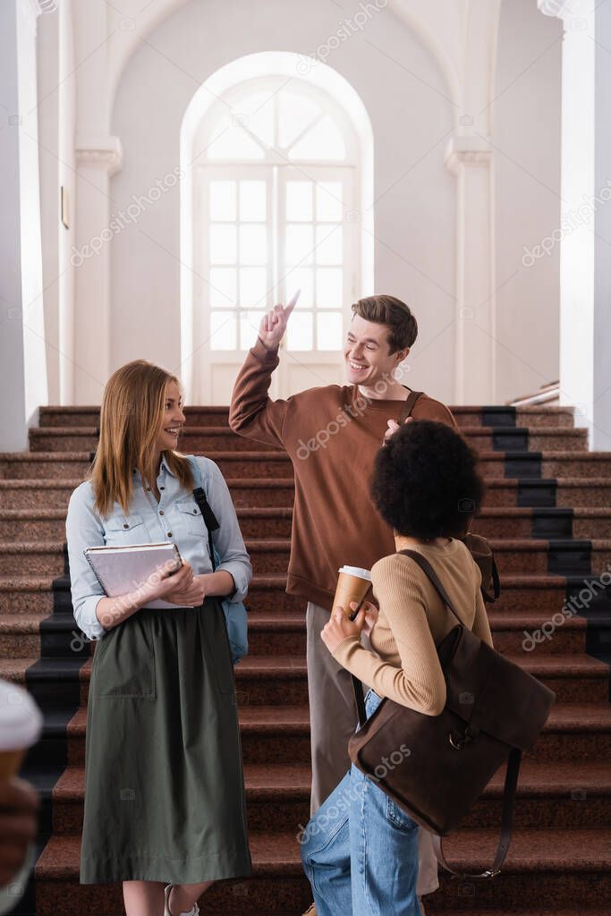 Smiling student pointing with finger near interracial friends in hall of university 