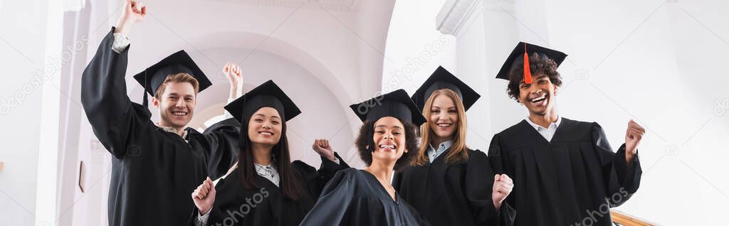 Cheerful multiethnic students in academic gowns showing yes gesture, banner 