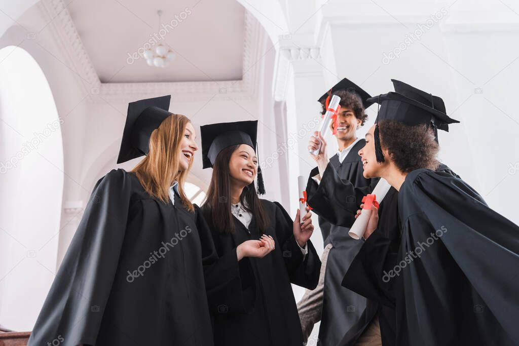 Low angle view of smiling interracial graduates with diplomas in university hall 