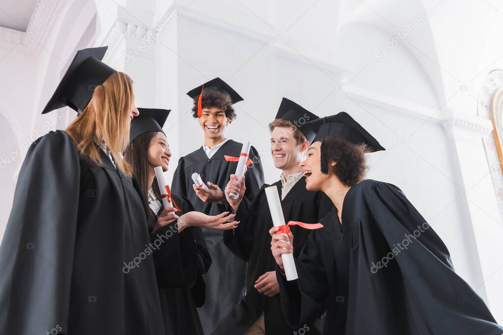 Cheerful interracial graduates in caps holding diplomas 