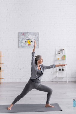 Savaşçı pozu veren spor giysili orta yaşlı kadın yoga yapıyor.