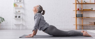 Kobra giymiş orta yaşlı bir kadın evde yoga yapıyor, afiş yapıyor.