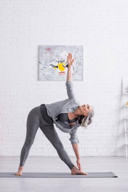 Orta yaşlı bir kadın üçgen duruşunda yoga yaparken evde poz veriyor. 