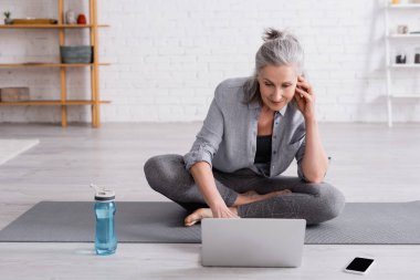 Nilüfer çiçekli orta yaşlı kadın yoga minderinde poz veriyor ve dizüstü bilgisayarda online özel ders izliyor. 