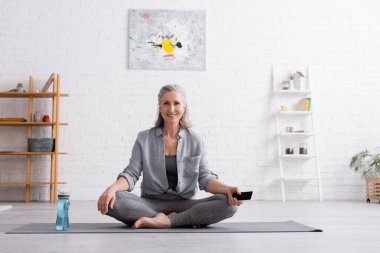 Nilüfer çiçeğinde oturan mutlu orta yaşlı kadın yoga minderinde poz veriyor ve elinde boş ekranla akıllı telefon tutuyor.