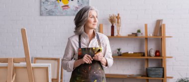 Tabelanın yanında fırça tutan önlüklü orta yaşlı bir sanatçı.