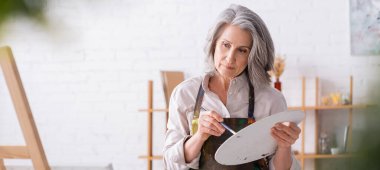 Önlüklü, fırça ve palet tutan düşünceli olgun bir kadın sehpaya, afişe bakarken