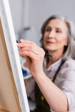 Bulanık ön planda resim yaparken elinde fırça tutan olgun bir kadın. 