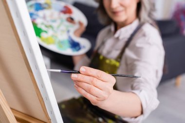 partial view of mature woman holding paintbrush while painting on canvas with blurred foreground  clipart